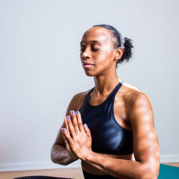 A woman wearing workout clothes sits in a yoga pose, with her eyes closed and her hands pressed together