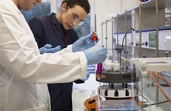 Two scientists, one in the foreground wearing a white lab coat and a second behind wearing a navy blue lab coat, looking at a purple liquid in a test tube