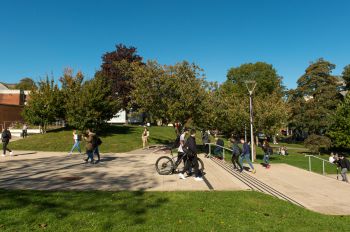 Various groups of people are walking across campus while pushing their bike and chatting to their friends.