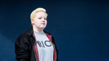 A young person with dyed blonde hair stands up wearing a dark jacket and light t-shirt with a blue background