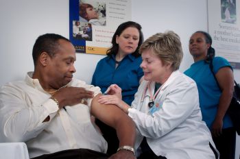 A man of colour rolls up a sleeve to receive a injection from a white female doctor while another healthcare staff member and an apparent family member look on
