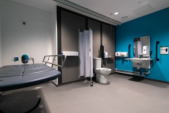 view of the changing place toilet including from left to right, a changing bench, shower, privacy screen, accessible wc and large basin with mirror. The walls are painted in white, grey and bright blue.