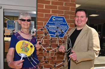 Lady Kroto and son Stephen hold a model of the buckyball and a yellow paper fan with the image of Prof Sir Harry Kroto on it. They stand in front of a newly unveiled blue plaque for Sir Harry Kroto.