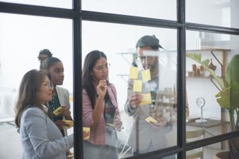 3 women and one man viewed through an office glass pane putting sticky notes on the glass