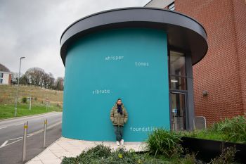 Helen Cammock stands in front of the artwork on an external wall at top of University of Sussex Student Centre. The text is painted in white and set against a bright teal background, and reads: whisper  tones  vibrate  foundations