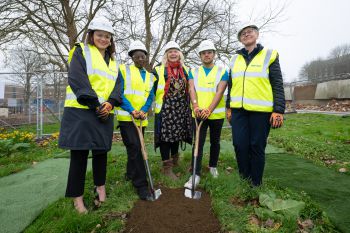 Five people wearing hard hats and high-vis jackets pose next to two spades and a freshly dug plot of earth.