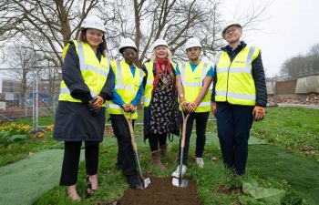Five people wearing hard hats and high-vis jackets pose next to two spades and a freshly dug plot of earth.