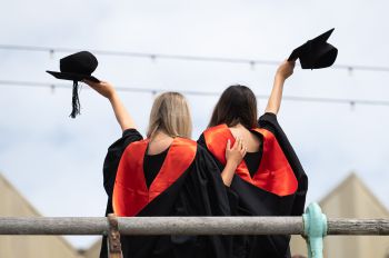 The backs of two graduates wearing graduation outfits with their arms around each other. They are posing on the Brighton beach front.