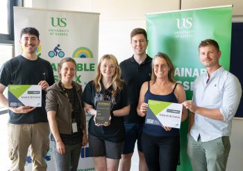 The Life Sciences Green Impact team standing together, smiling and holding their awards in front of sustainability banners
