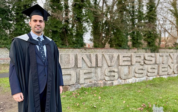 A student in graduation robes standing by the University of Sussex concrete sign