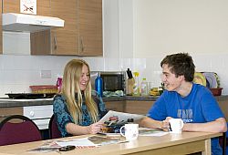 A female student reading a paper at the table of a kitchen in Stanmer Court. A male student sits beside her with a cup of coffee