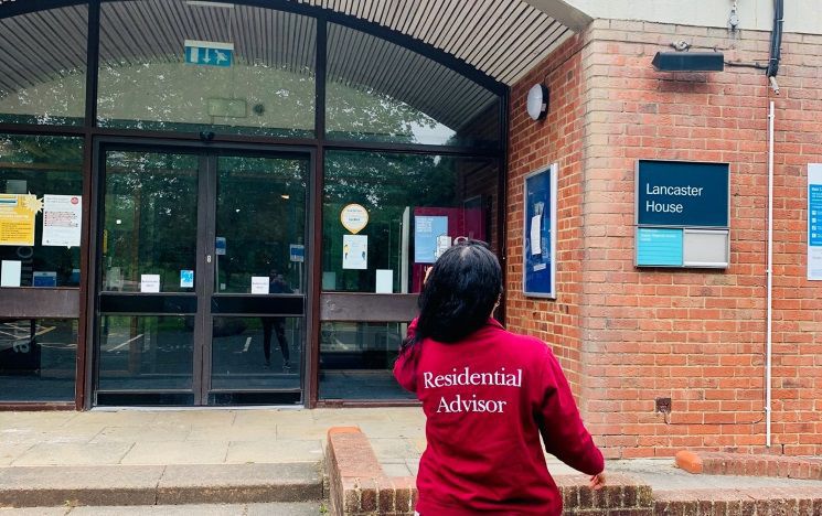 A student faces away from the camera and towards a red brick university building wearing a Residential Advisor hoodie