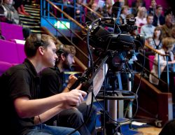 A man uses a television camera while, in the background, audience members sit in raked seating