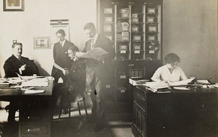 Black and white image of Eva Sommer working at a desk on the right hand side of the office with four male workers on the left hand side.
