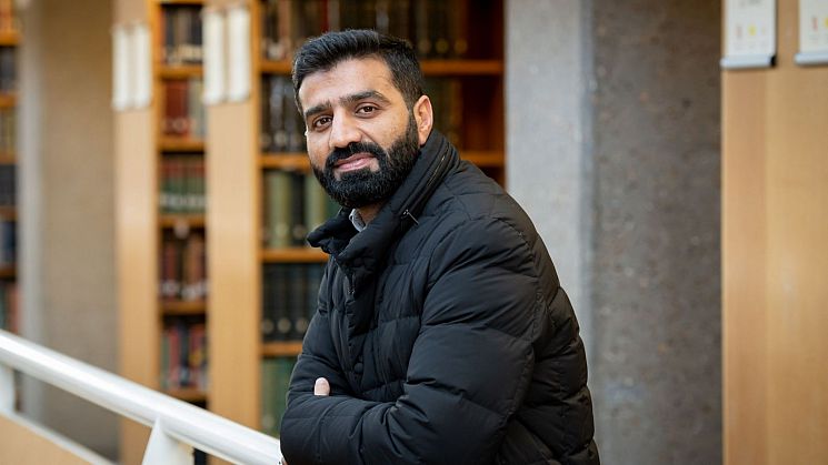 Naimat standing in the university library with bookshelves behind him