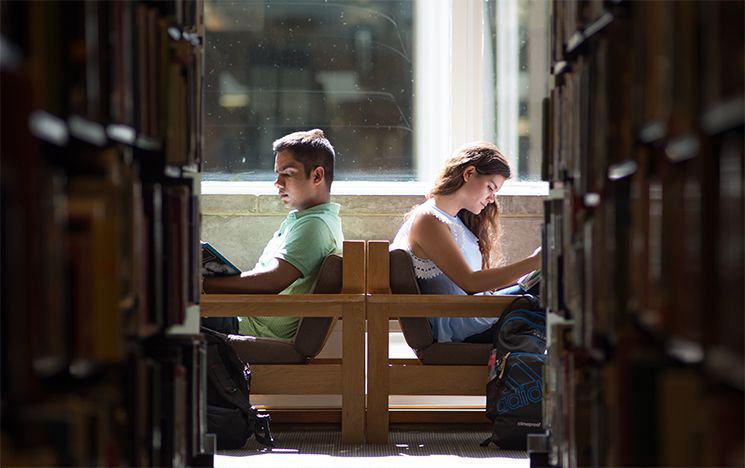 Two students sat in back-to-back chairs at the end of an aisle of books in the library