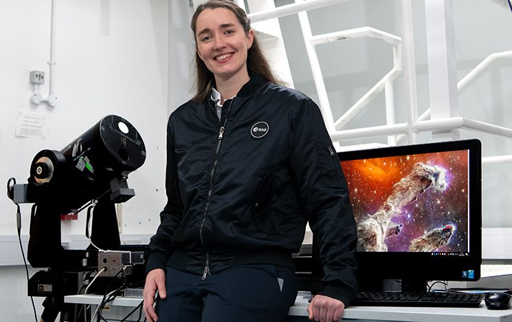 Dr Rosemary Coogan perches on desk in the Astronomy lab, with telescope and monitor showing pillars of creation JWST image