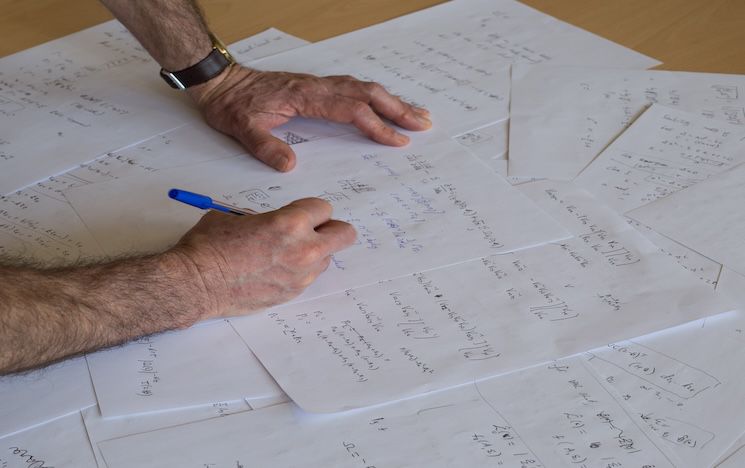 Table covered in Maths calculations with two hands - one holding a pen.