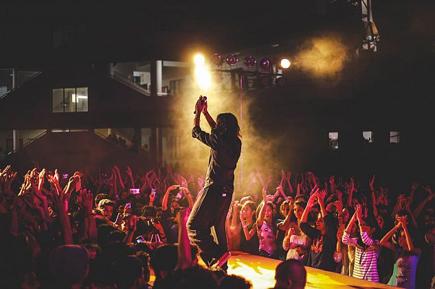 image of a singer on stage clapping their hands - they are surrounded by a crowd who also have their hands clapping about their heads