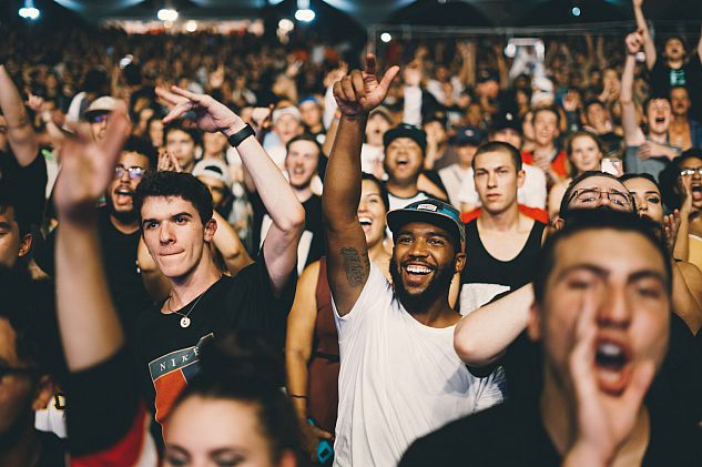image of a crowd at a music concert. two men are in focus in the middle of the image, both with one hand in the air. one purses his lips, the other smiles widely