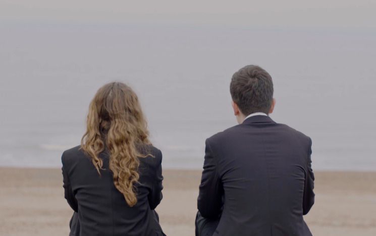 Photograph of man and woman taken from behind (only shows their backs) sitting on a beach looking at the water
