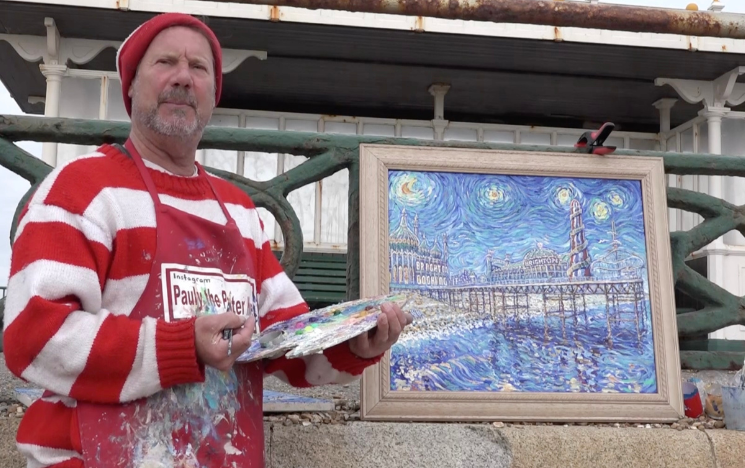 A man in a red jumper and skull cap stands next to his art work in an outdoor setting. It is a painting of Brighton, depicting the Royal Pavilion and the pier, with inspiration from renowned artist Vincent Van Gogh.