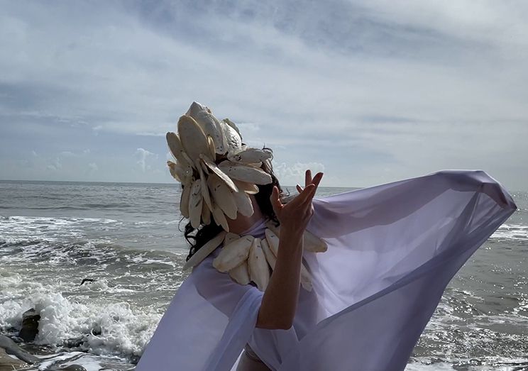 Photo of a white person swear a mask make out of shells and wearing purple fabric, against a backdrop of the sea.