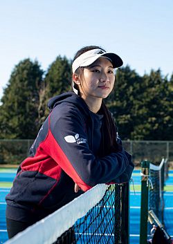 A female student bending over a tennis net in the sun with a tennis racket in her hand