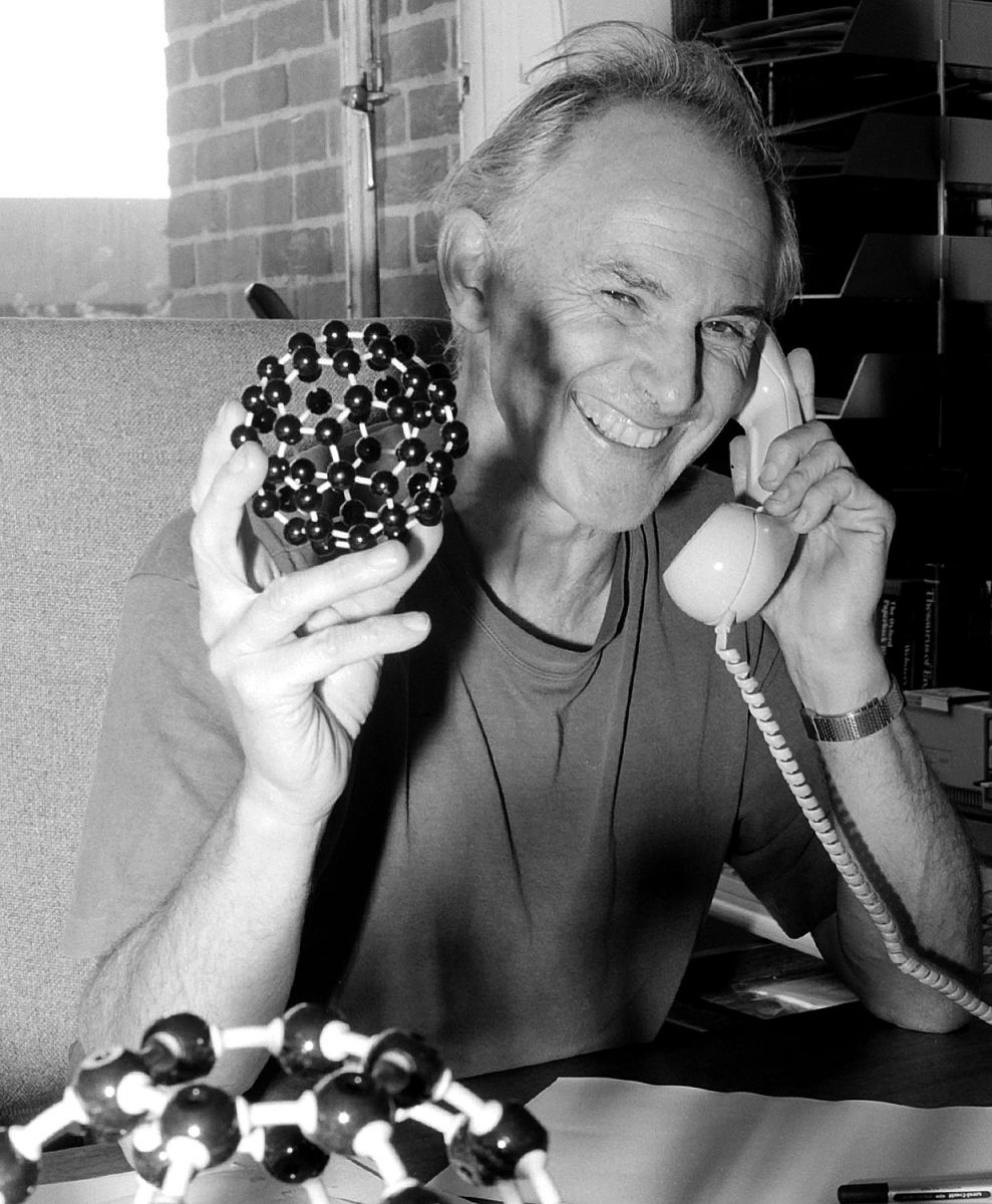 Black and white archive photograph of Harry Kroto holding up a model buckyball while using the telephone.