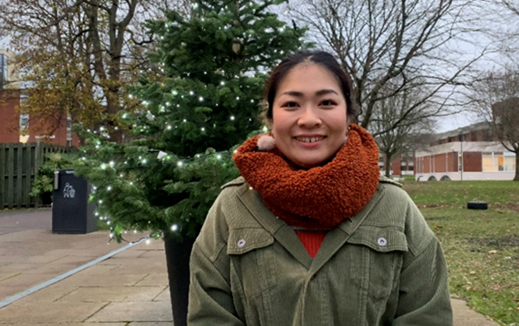 Student stands in front of a Chrsitmas tree on campus smiling on an overcast day