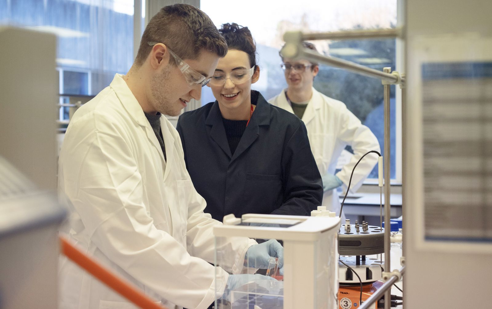Male and female student in a lab, goggles on, handling test tubes