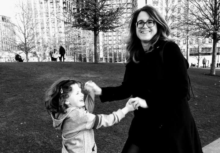  A black and white photo of Sarah-Jane and her daughter. They're holding hands and dancing outside on a wall, by a grass area in a city