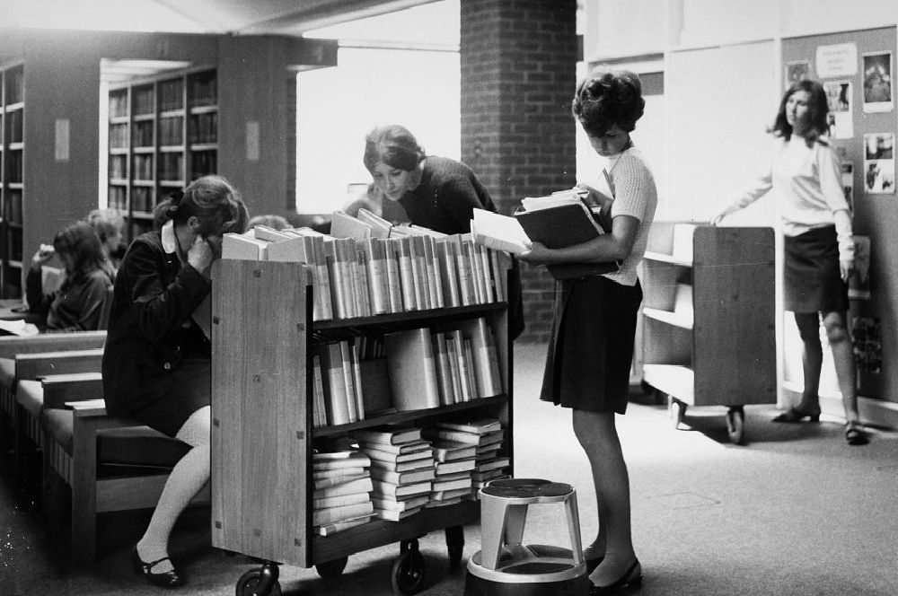Black and white photo of female students in the Library, wearing 60s-style clothes and hair.