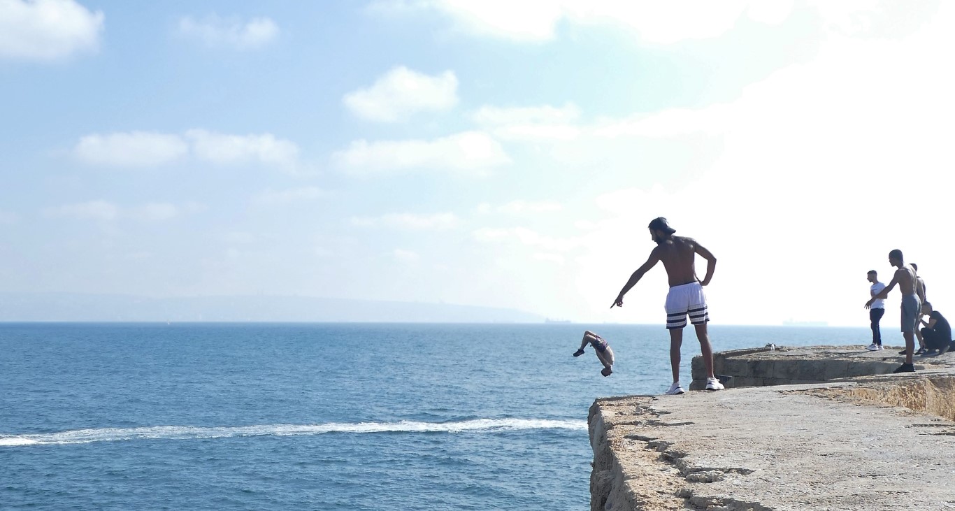 
Young men jump off of a rocky cliff into the sea on a sunny day