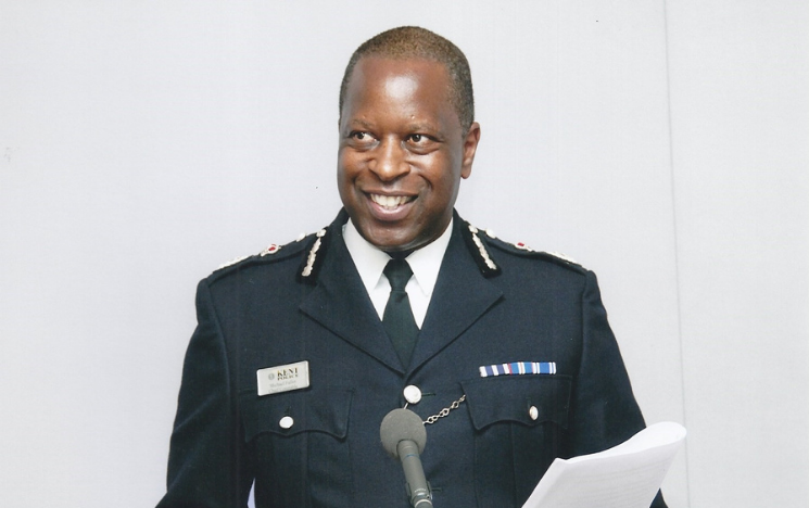 A photo of Michael Fuller in uniform at a lectern, smiling at someone off-camera as he reads from a sheet of paper