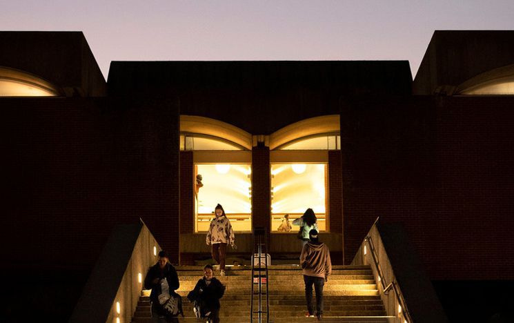 A photo of the outside of the library at night time, with students walking up the stairs and the entrance lit up