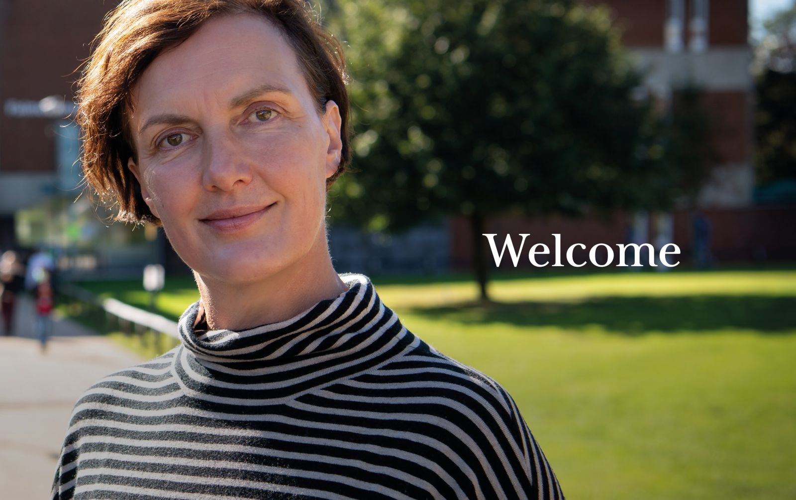 A photo of the Provost in front of Falmer House, smiling at the camera on a sunny day beside the word 'Welcome'