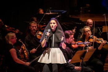 Music artist Gazelle Twin stands in a spotlight on stage holding a mic, wearing a black hooded cape and grey skirt. Behind her, dimly lit, are members of a string orchestra.