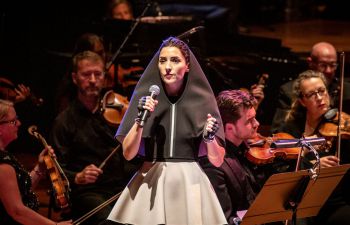 Music artist Gazelle Twin stands in a spotlight on stage holding a mic, wearing a black hooded cape and grey skirt. Behind her, dimly lit, are members of a string orchestra.