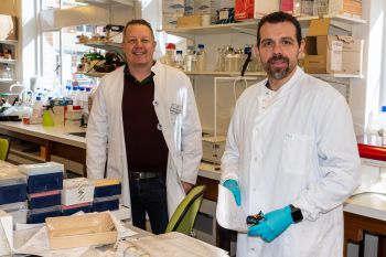 Two scientists in white coats stand side-by-side in a lab facing the camera