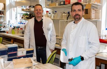 Two scientists in white coats stand side-by-side in a lab facing the camera