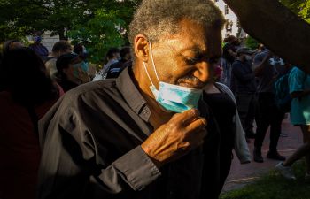 A black man in a black shirt stands aside from a crowd in the shade of a tree