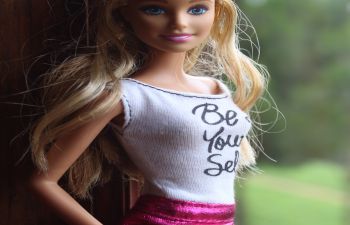 A Barbie doll stands by a window wearing a pink skirt and white top with the text Be Your Self written on it