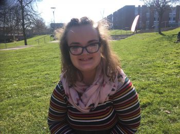 Emma Beeden sits on a grass slope in a stripey jumper with the bright sun in the background
