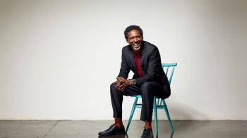Lemn Sissay MBE is photographed sitting on a white chair in a studio. He is dressed smartly in a jacket and smiling at the camera