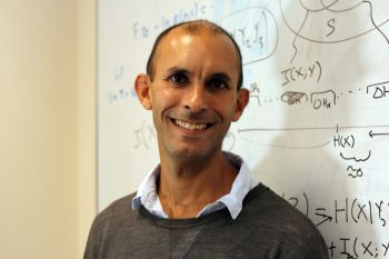 Professor Anil Seth stands in front of a white board with mathematical equations written in black pen wearing a grey jumper and blue shirt