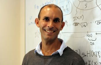 Professor Anil Seth stands in front of a white board with mathematical equations written in black pen wearing a grey jumper and blue shirt