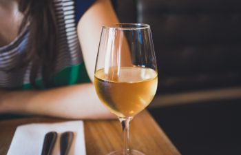 A close up shot of a glass of white wine with a woman wearing a blue and white stripey top in the background sitting at a restaurant table