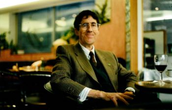 Dr (Jim) Perry James Browne seated by a table with a glass of red wine on it. He is smiling and wears glasses.