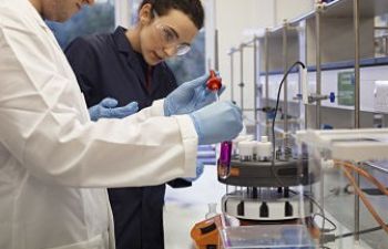 Two scientists, one in the foreground wearing a white lab coat and a second behind wearing a navy blue lab coat, looking at a purple liquid in a test tube